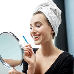 A woman holds a syringe of bleaching gel and a mirror while sitting at home and whitening her teeth